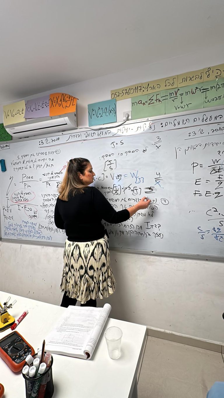 חנה קדמי עומדת וכותבת על לוח מחיק בחדר שמצויד עם כל הציוד המשוכלל לצילום ולעריכת שיעור.